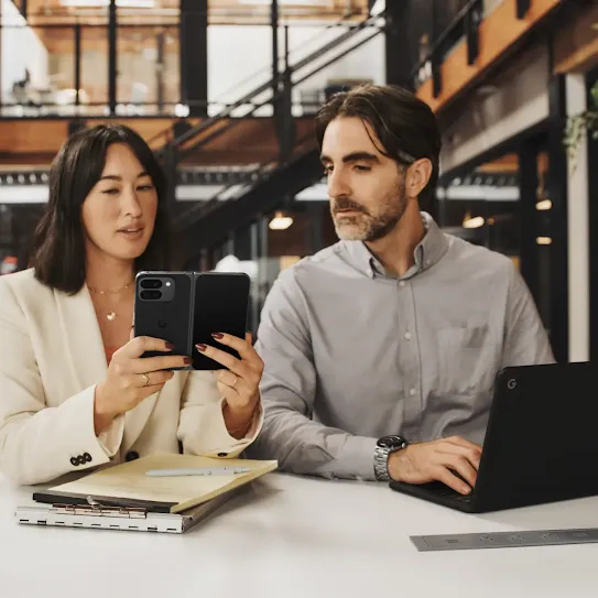 Two people seating at a desk in a modern office, looking at the phone together while working on laptops and notebooks.