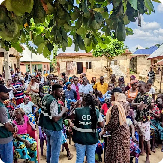 Field workers wearing green GiveDirectly vests speak to a large crowd gathered outdoors in a village setting during a community outreach or distribution meeting.