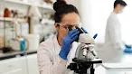 A female scientist wearing safety goggles, blue gloves, and a white lab coat leans forward to look through a microscope in a brightly lit laboratory.