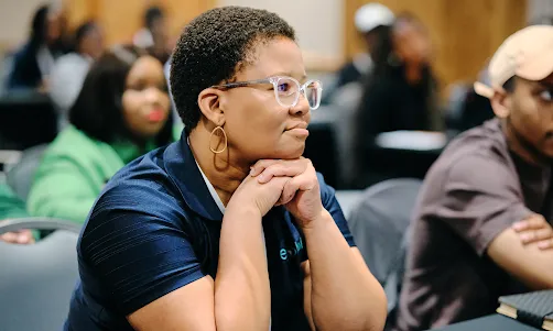 A Black woman with short hair and clear glasses sits attentively at a conference, resting her chin on her clasped hands.