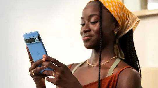 Una mujer con un pañuelo amarillo estampado sonríe mientras usa un smartphone Google Pixel azul para enviar un mensaje.