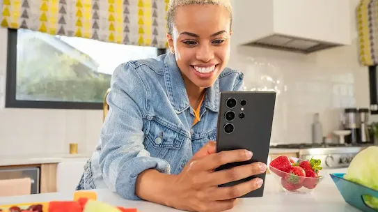 Eine lächelnde junge Frau verwendet ein schwarzes Smartphone in einer hellen Küche, in der frisches Obst auf der Theke steht.