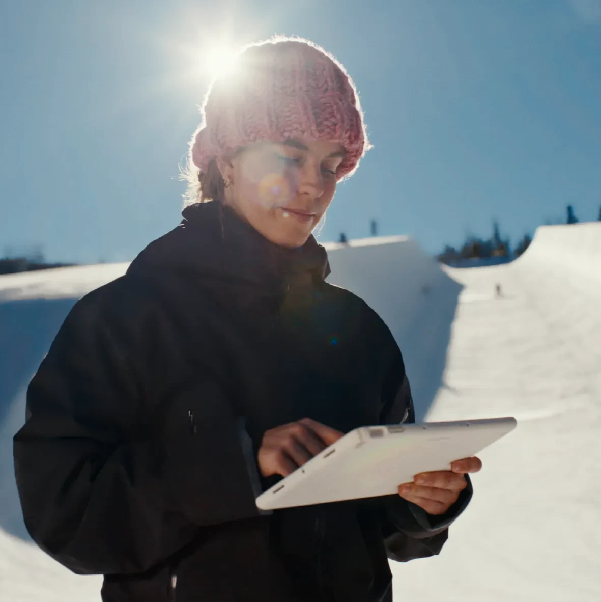 A woman outdoors in the snow looks at a tablet. A half pipe is behind her.