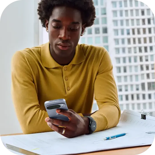 Um homem sorri sentado em um sofá ao ar livre, olhando para seu smartphone Pixel azul-claro e usando um smartwatch para combinar.