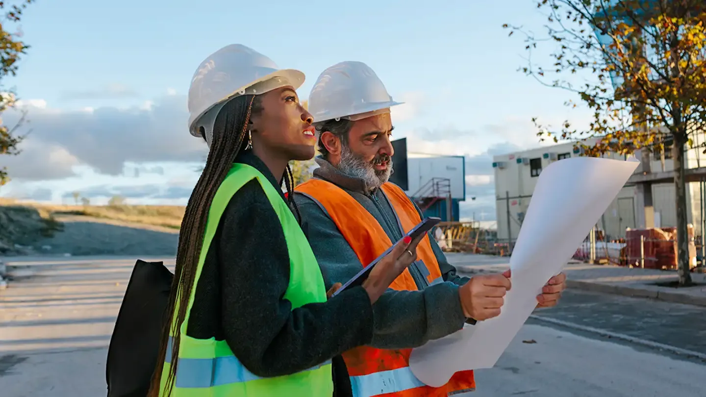 A Black woman wearing a hard hat and a yellow safety vest inspecting an outdoor job site using a mobile tablet with a Middle Eastern male colleague wearing a hard hat and an orange safety vest.