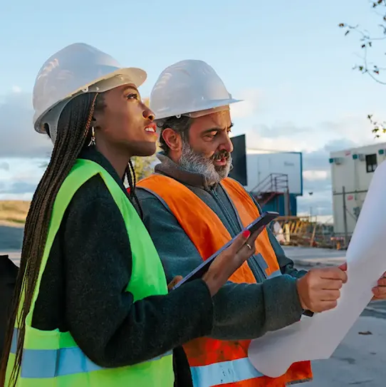 A female and male engineer wearing white hard hats and safety vests standing outdoors, reviewing architectural blueprints together at a construction site.