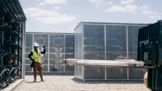 A worker guides the installation of a new component into an outdoor EV battery center.
