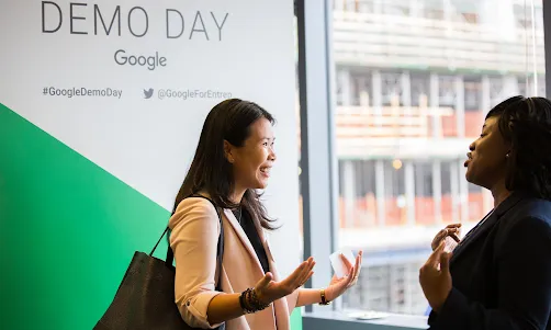 A woman wearing blue top in focus talking to another woman facing away from the camera.