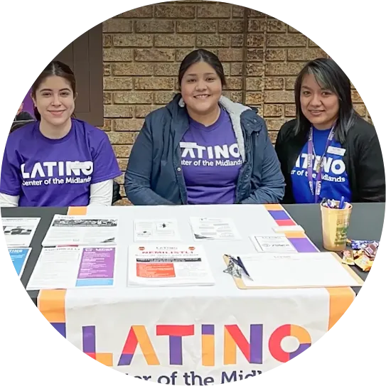 Three women in purple "Latino Center of the Midlands" t-shirts smile behind an information table with brochures and sign-up sheets, framed within a circular crop.