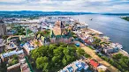 An aerial view of a historic city featuring a massive, castle-like building with a green roof situated on a hill overlooking a wide river and waterfront docks.