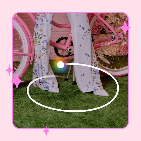A close up of Paris Hilton’s floral print pants and white pointed, standing on green turf in front of a pink bicycle. A white oval illustrating an example of the Circle to Search feature, is drawn in front of the shoes.