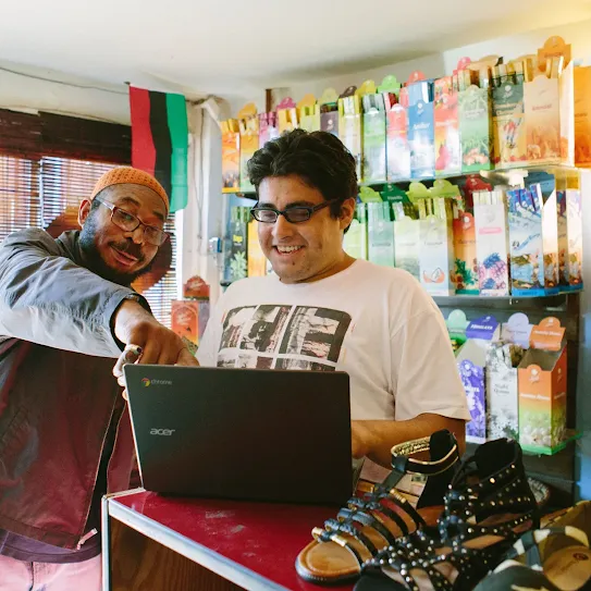 Two men inside a brightly lit retail shop look together at a black Acer Chromebook laptop resting on a counter. The man on the left, wearing a brown jacket, glasses, and an orange knit cap, leans in and points at the screen. The man on the right, wearing glasses and a graphic t-shirt, smiles as he works on the computer. Shelves behind them are filled with colorful packaged goods, and sandals are displayed on the counter in the foreground.
