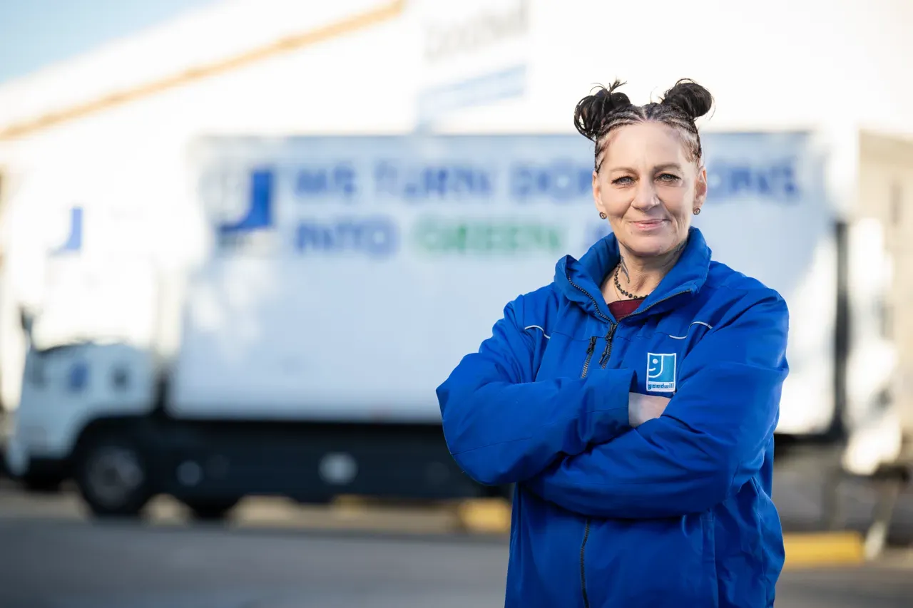 A Goodwill employee wearing a blue logo jacket stands with her arms crossed, smiling in front of a white Goodwill donation truck. The image represents Goodwill's commitment to providing AI training at scale across its vast network of local community centers.
