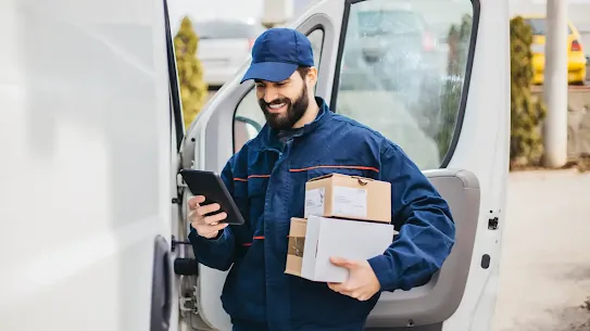 A smiling delivery driver in a blue uniform and cap holding several packages and checking a handheld tablet in front a white delivery van.