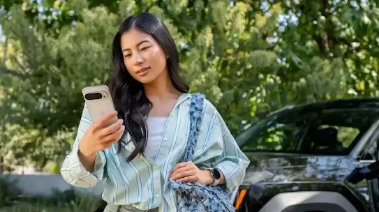 Uma mulher parada numa área externa com um carro ao fundo. Ela está segurando um smartphone branco e uma bolsa azul e branca com estampas.