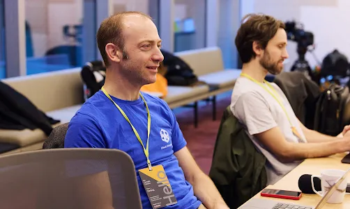 Two people are sitting side-by-side. One person is wearing a blue t-shirt and has an ID card hanging around their neck.
