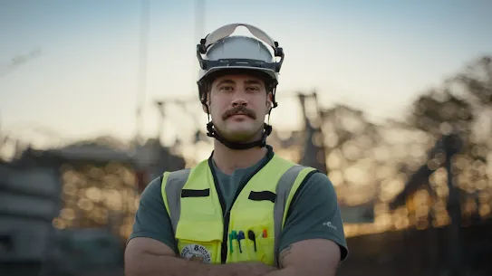 A medium close-up shot of a male worker with a mustache, standing with his arms crossed and looking directly at the camera. He is wearing a white safety hard hat with a clear visor pushed up, a high-visibility neon yellow safety vest with reflective strips, and a dark green t-shirt. Pens are tucked into the pocket of his vest. The background is softly blurred, showing an outdoor industrial or construction site with structures and trees silhouetted against a golden hour sky.