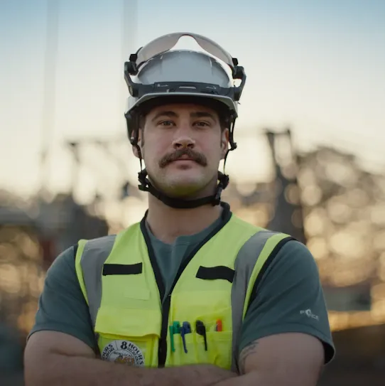 A medium close-up shot of a male worker with a mustache, standing with his arms crossed and looking directly at the camera. He is wearing a white safety hard hat with a clear visor pushed up, a high-visibility neon yellow safety vest with reflective strips, and a dark green t-shirt. Pens are tucked into the pocket of his vest. The background is softly blurred, showing an outdoor industrial or construction site with structures and trees silhouetted against a golden hour sky.