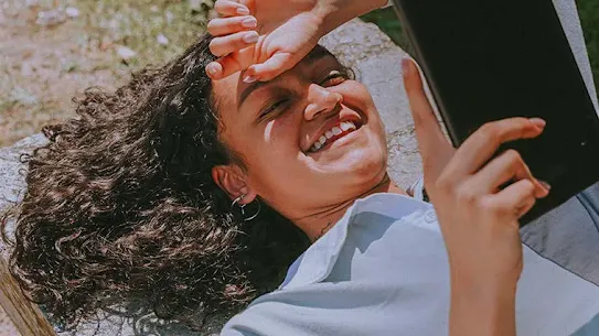 A smiling young woman with curly hair lying outdoors on a stone bench, holding a tablet while shielding her eyes from the sun.