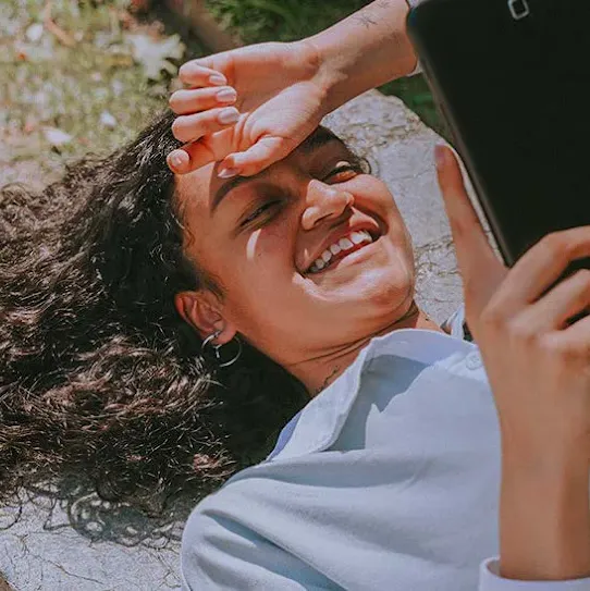 A smiling young woman with curly hair lying outdoors on a stone bench, holding a tablet while shielding her eyes from the sun.