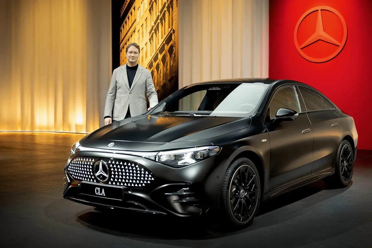 Ola Källenius, CEO of Mercedes-Benz, standing next to a sleek, matte black Mercedes-Benz CLA concept car on a stage. The background features a large red wall with the Mercedes-Benz logo and warm stage lighting.