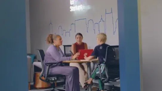 three women sitting around a table with a laptop talking.