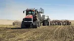 A large, heavy-duty red tractor with track treads pulls a massive seeding or tilling implement across a dry, expansive agricultural field under a clear blue sky.