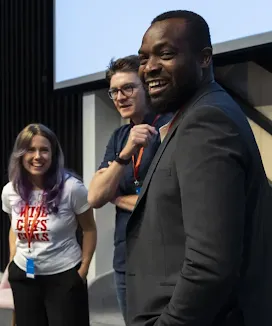 Tres oradores de pie sobre un escenario en un evento, sonriendo e interactuando con el público durante una presentación.