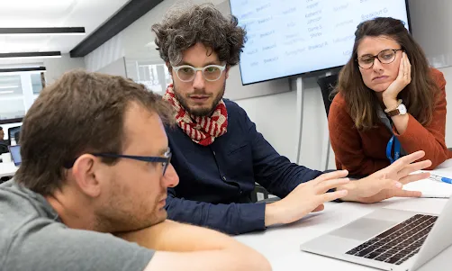 Three individuals are discussing something with a laptop on the table.