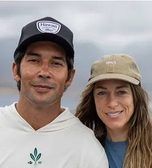 A smiling man and woman wearing baseball caps (one labeled 'Hawaii' and the other 'Maui') posing outdoors against a soft, blurred coastal background.