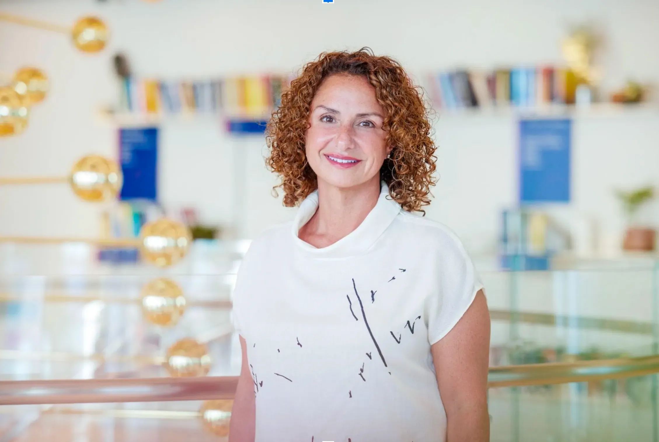 Professional headshot of Lila Ibrahim, Chief Operating Officer at Google DeepMind. She is smiling and wearing a white top, standing in a bright office space with bookshelves and decorative gold spheres in the background.