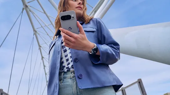 A person wearing a blue leather jacket and jeans is holding a Moonstone Google Pixel 10 phone underneath a structure outside.
