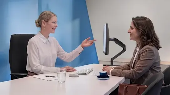Two women sitting at an office desk, with one pointing at a computer monitor while talking to the other.