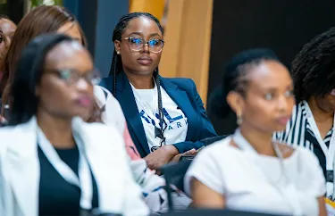 A professional woman with braided hair and glasses sits attentively in an audience at a corporate event or conference.