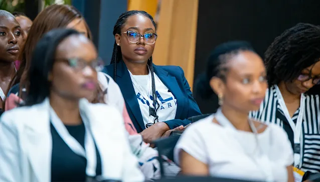 A professional woman with braided hair and glasses sits attentively in an audience at a corporate event or conference.