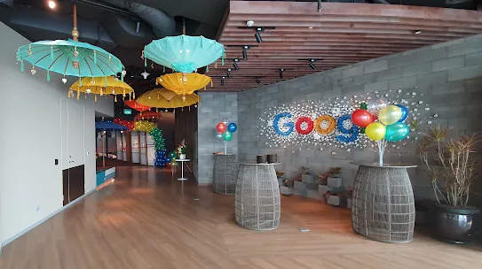 A Google office space decorated for a celebration. Groups of colorful balloons sit on wireframe tables and large, silk umbrellas hang from the ceiling.
