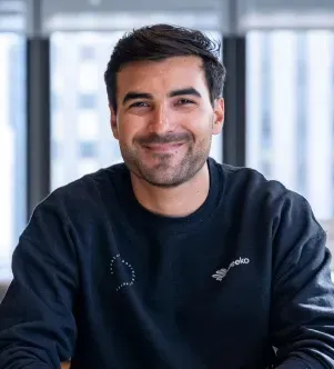 A smiling man with dark hair and a short beard, wearing a black sweatshirt, sitting in a bright office with large windows and city buildings in the blurred background.