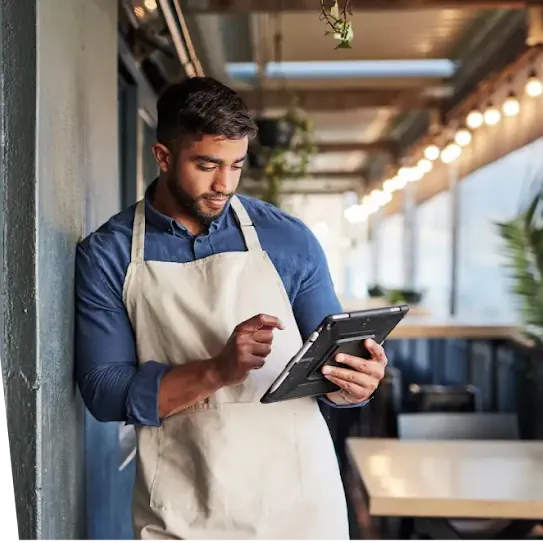 A man wearing a beige apron and blue shirt leans against a wall while using a digital tablet in an outdoor café setting.
