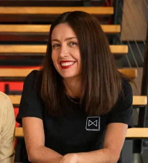 A professional portrait of Adriana Cisneros Basulto, co-founder of Maxwell, smiling in a black t-shirt while sitting on indoor stairs, featured in a Nebraska case study on AI-personalized HR benefits.