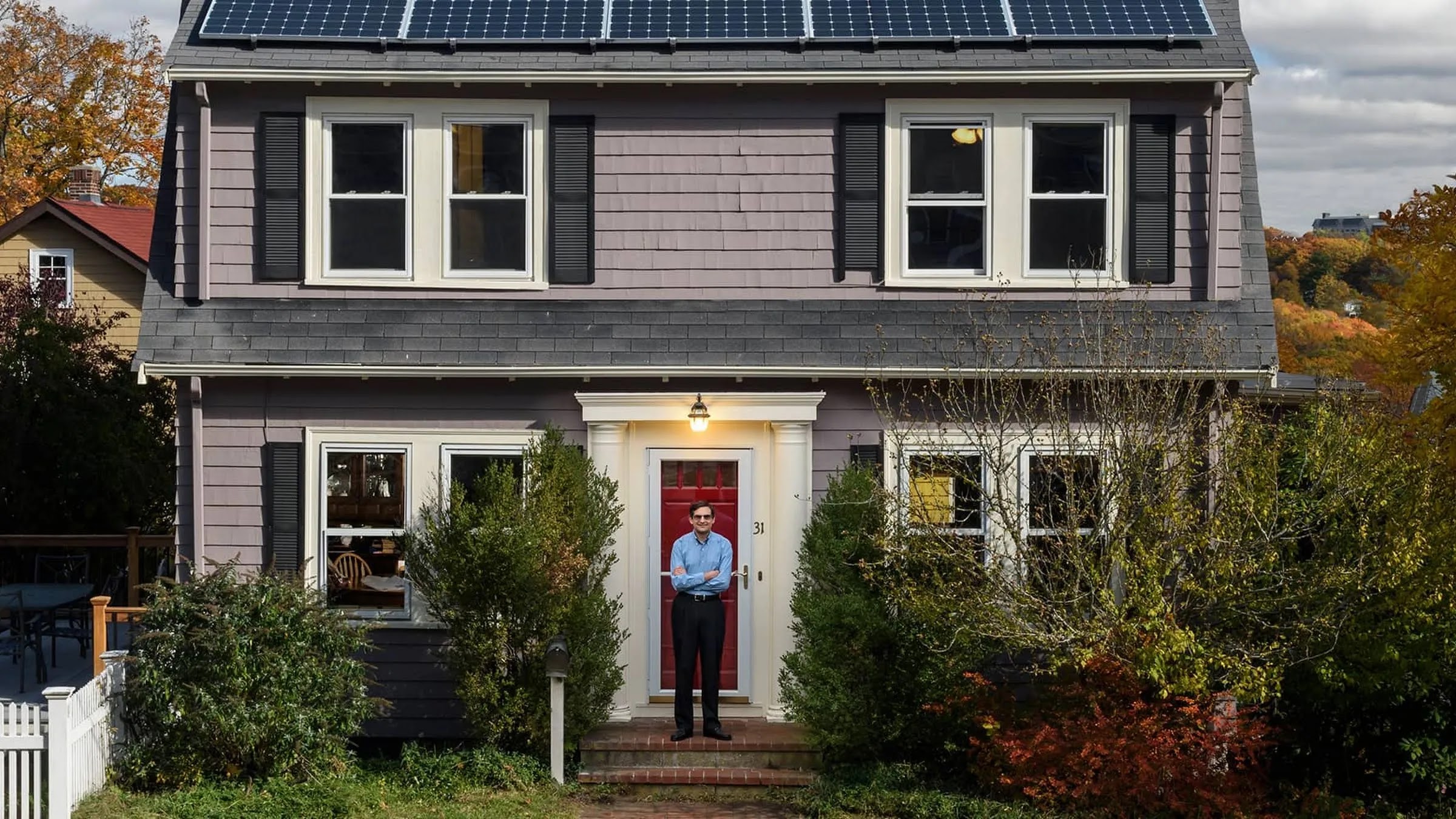 A man stands in front of a quaint home with solar panels on the roof.