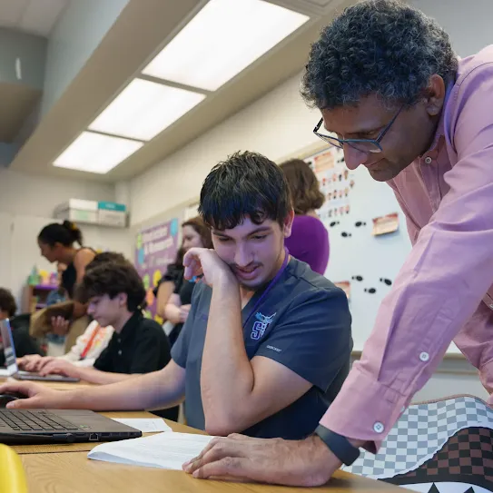 Male teacher reading a page with student looking at same paper at desk in a class room full of students and faculty 
