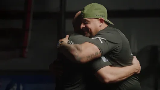 Two male veterans in olive green t-shirts sharing a supportive embrace in a dimly lit gym environment. This image highlights the community aspect of the Google.org Fellowship and ReflexAI's mission to support veteran mental health.
