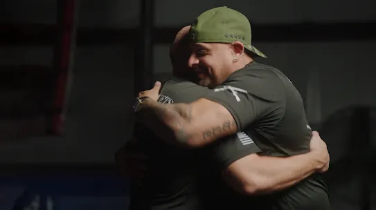 Two male veterans in olive green t-shirts sharing a supportive embrace in a dimly lit gym environment. This image highlights the community aspect of the Google.org Fellowship and ReflexAI's mission to support veteran mental health.