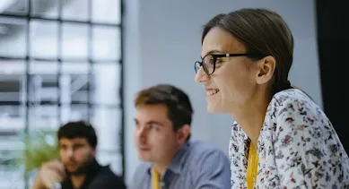 Woman wearing glasses is looking away from the camera and smiling. Two men are in the background in soft focus.