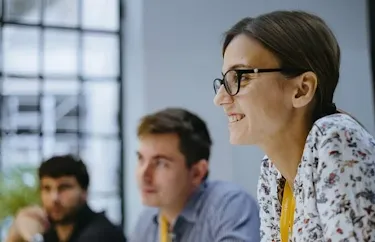 Woman wearing glasses is looking away from the camera and smiling. Two men are in the background in soft focus.