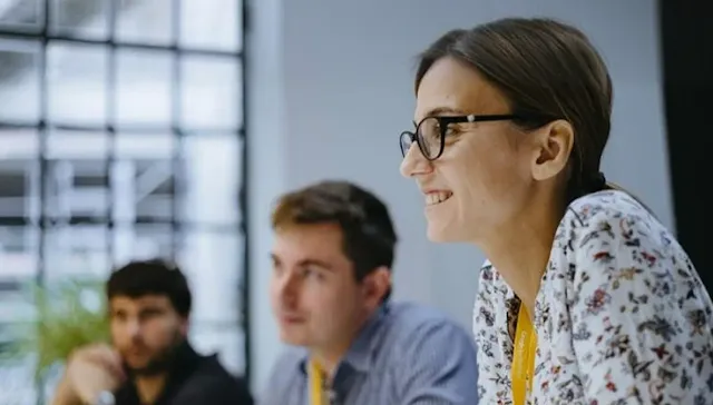 Woman wearing glasses is looking away from the camera and smiling. Two men are in the background in soft focus.