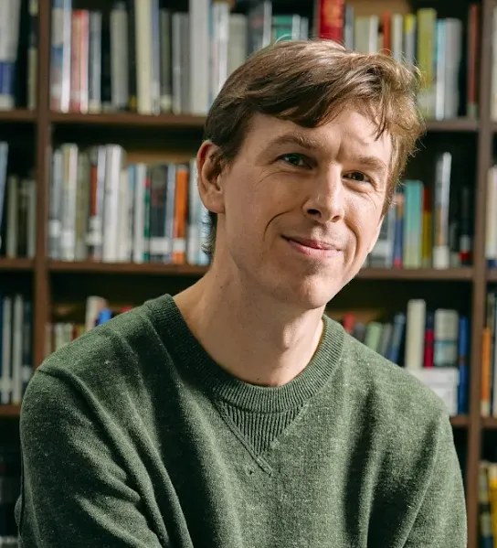 A portrait of a smiling man with light brown hair wearing a green textured sweater, sitting in front of a bookshelf filled with books, featured in a New York case study on AI in education.
