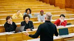 A professor in the foreground gestures toward a diverse group of university students seated at long wooden desks in a tiered lecture hall.