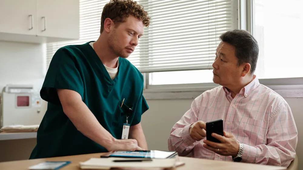 A male nurse in green scrubs stands at a table discussing health information on a tablet with an older Asian male patient holding a smartphone in a medical office.