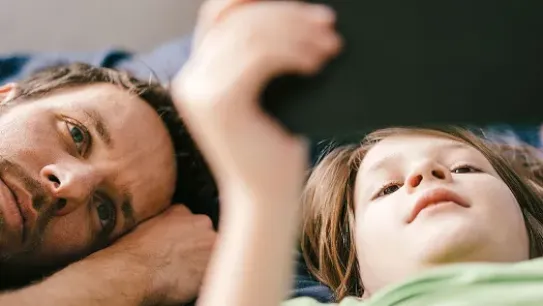 A close-up, shallow-depth-of-field photo of a father and young child lying down together. The child is holding a tablet (blurred in the foreground), while the father lies beside them, looking at the screen with an attentive and caring expression. The image conveys a sense of supervised digital exploration and youth safety.
